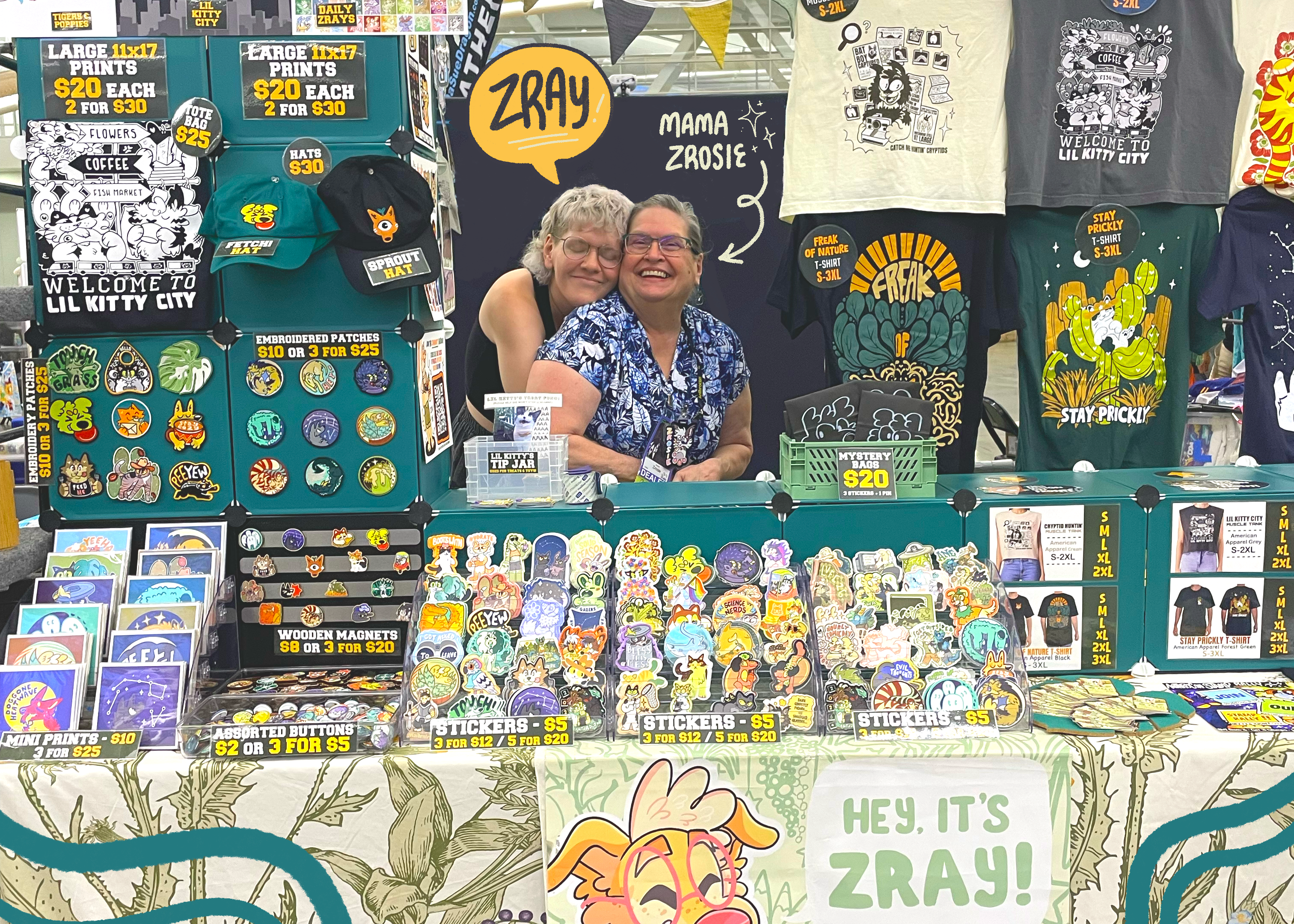 A colorful artist booth at a craft fair featuring merchandise from Hey,it’s Zray! The display includes graphic t-shirts, stickers, magnets, and art prints arranged on tables and hanging racks, with two people smiling behind the counter.